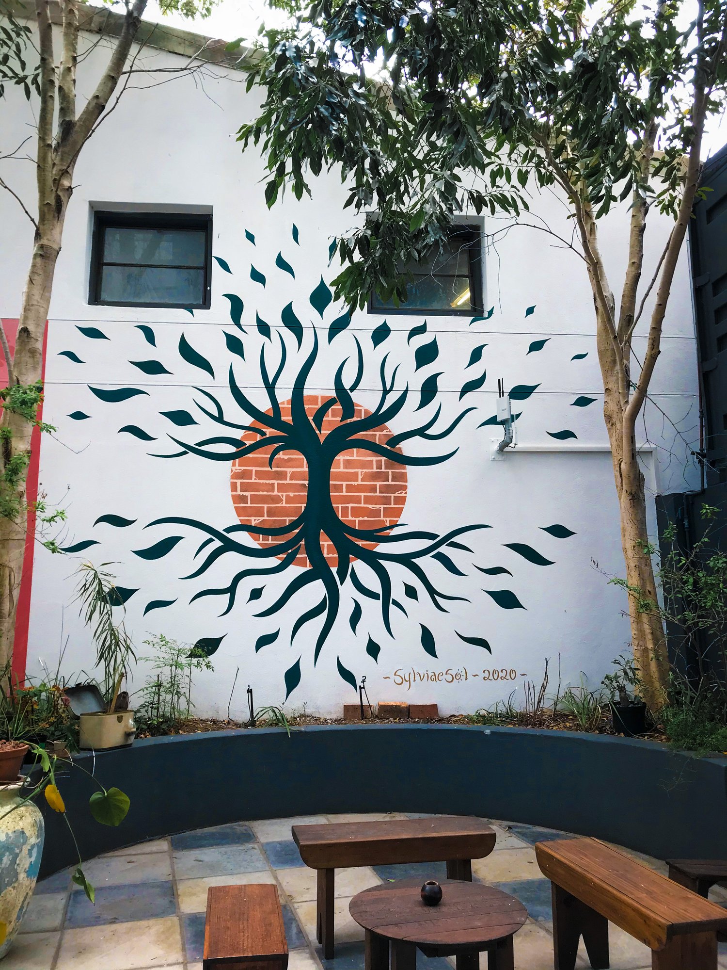Tree-of-life mural on a white courtyard wall, branches and roots radiating from a brick-circle sun, with cafe tables below.