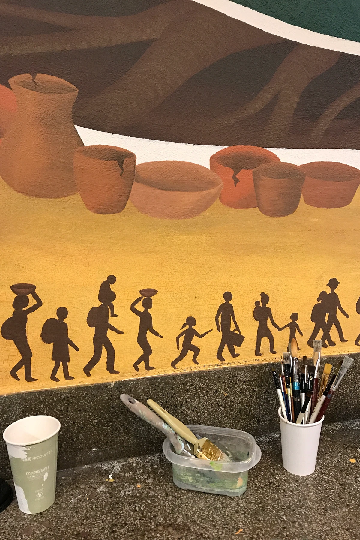 Pachamama mural detail: clay vessels above a row of silhouetted figures, with the artist’s brushes in the foreground.